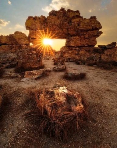 Lever de soleil à travers une formation rocheuse avec des herbes sèches au premier plan.